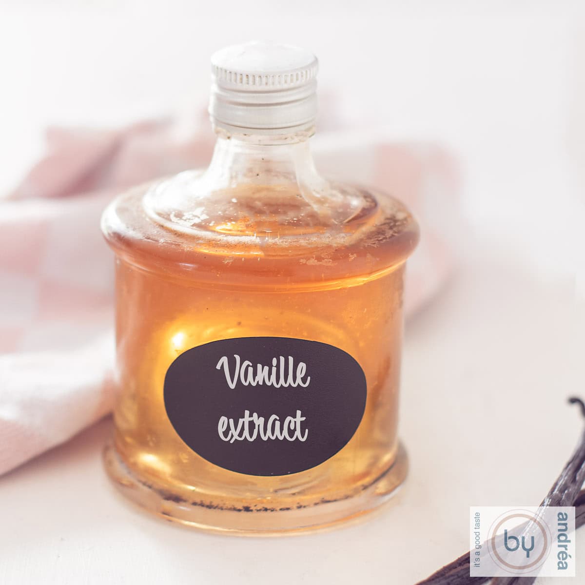 Een vierkante foto met een fles gevuld met vanille extract. Erachter een roze witte theedoek