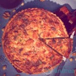 Een vierkante foto van een quiche waar een punt uit is gesneden. Een blauwe ondergrond waar de quiche op ligt