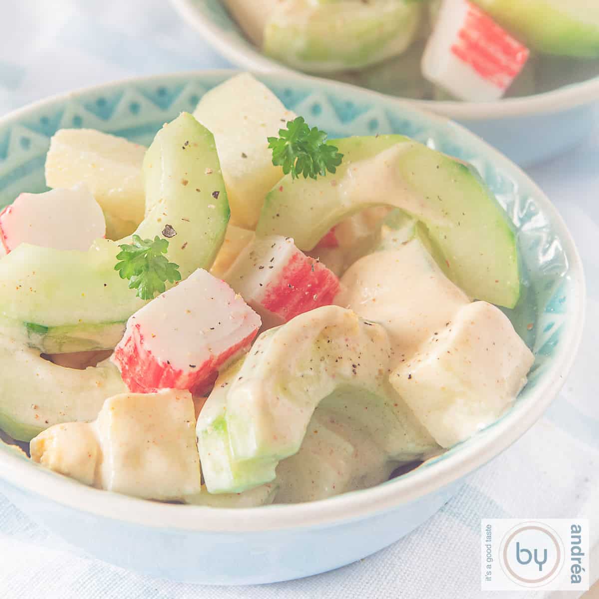 Een vierkante foto van een blauw schaaltje gevuld met komkommer, surimi, appel en ei salade.