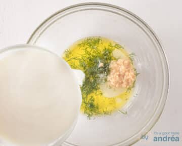 Een glazen schaal met olijfolie, zout, dille en knoflook. Er wordt Griekse yoghurt bijgeschonken.