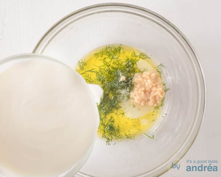 Een glazen schaal met olijfolie, zout, dille en knoflook. Er wordt Griekse yoghurt bijgeschonken.