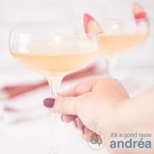 Een vierkante foto met twee glazen gevuld met champagne en rababer siroop. Een hand pakt het glas