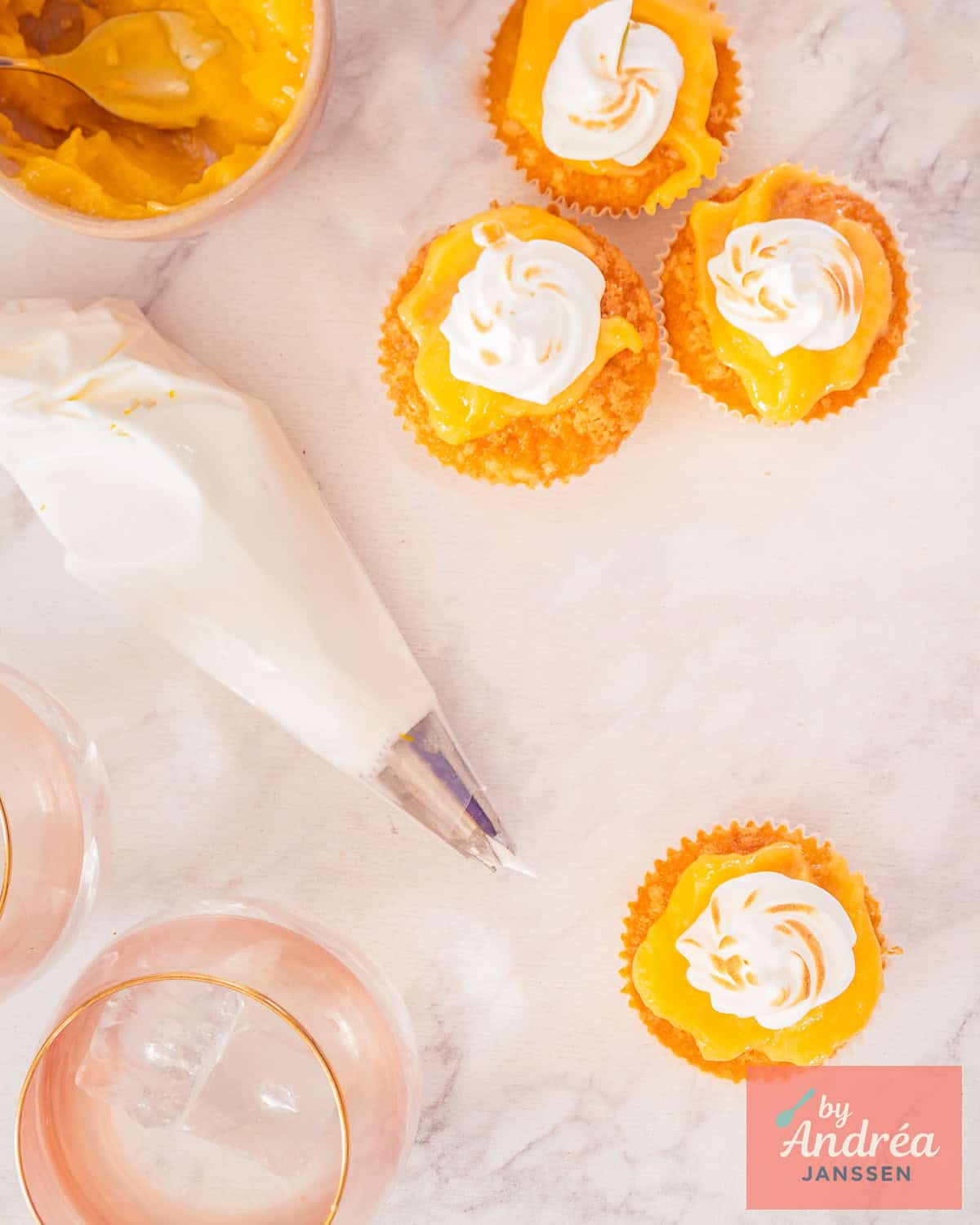 Een foto van bovenaf van een wit marmeren ondergrond met spuitzak vol meringue en vier citroen meringue cupcakes