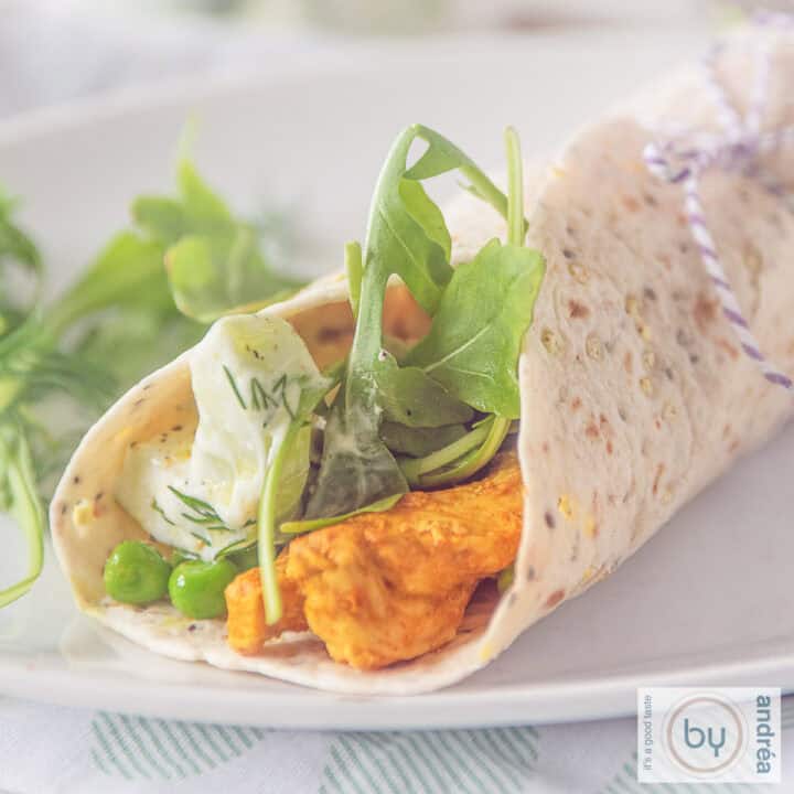 Een vierkante foto met een wit bord met een opgerolde wrap gevuld met Tandoori kip, tzatziki en groenten