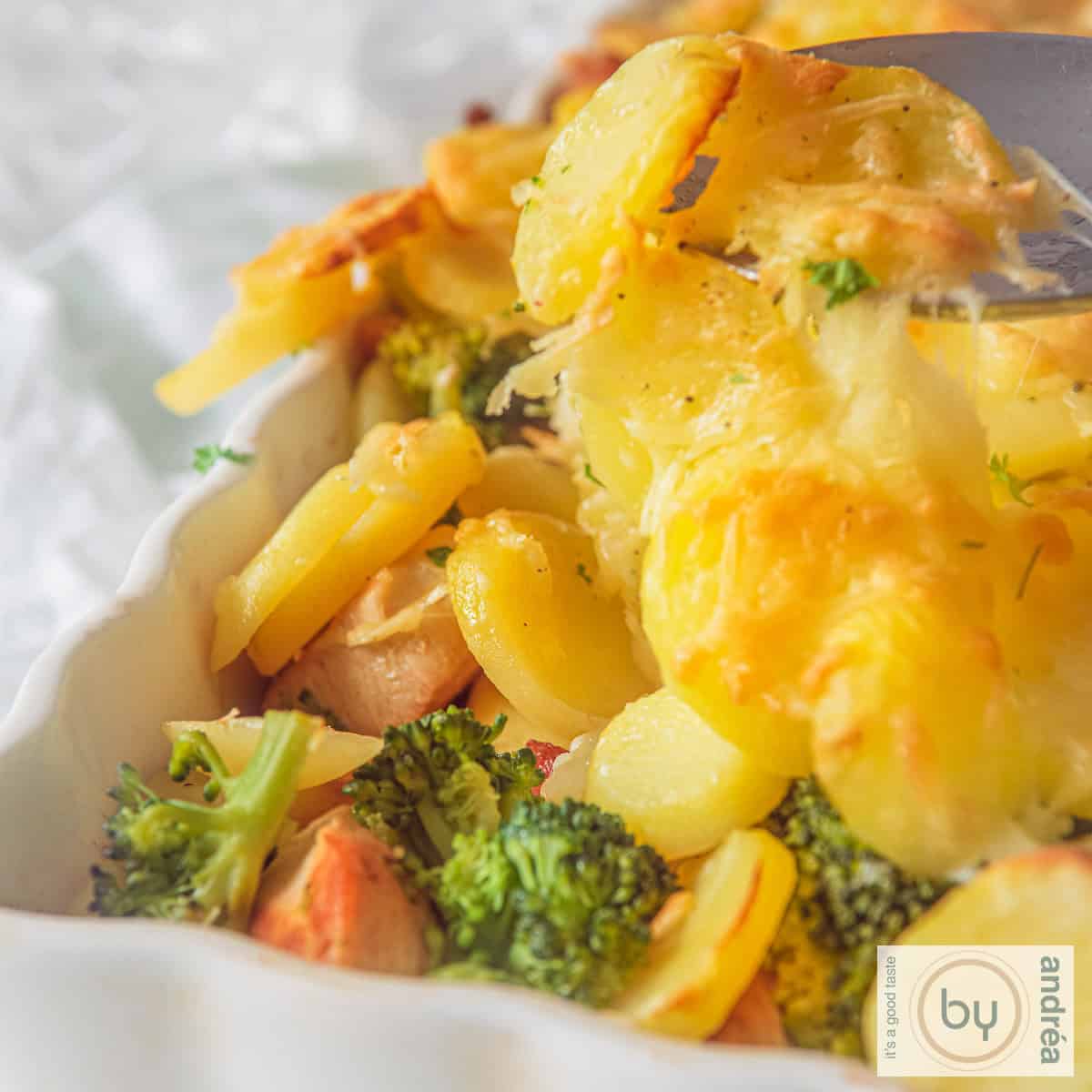 Een vierkante foto met een deel van een witte ovenschaal. Een bovenlaag van gegratineerde aardappels. Daaronder zie je broccoli en kip.