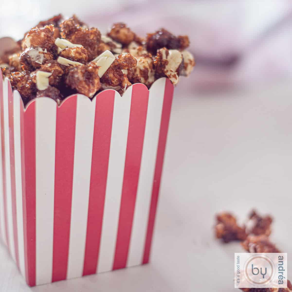 Een vierkante foto met een rood wit gestreept bakje gevuld tot de rand met karamel popcorn met witte chocola