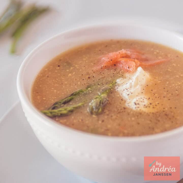 Een vierkante foto met een witte kom met Mosterdsoep met groene asperges en zalm