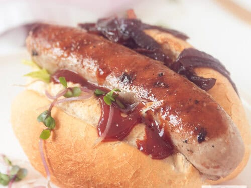 Een vierkante foto met een broodje gevuld met gegrilde worst en zoete gekarameliseerde uitjes