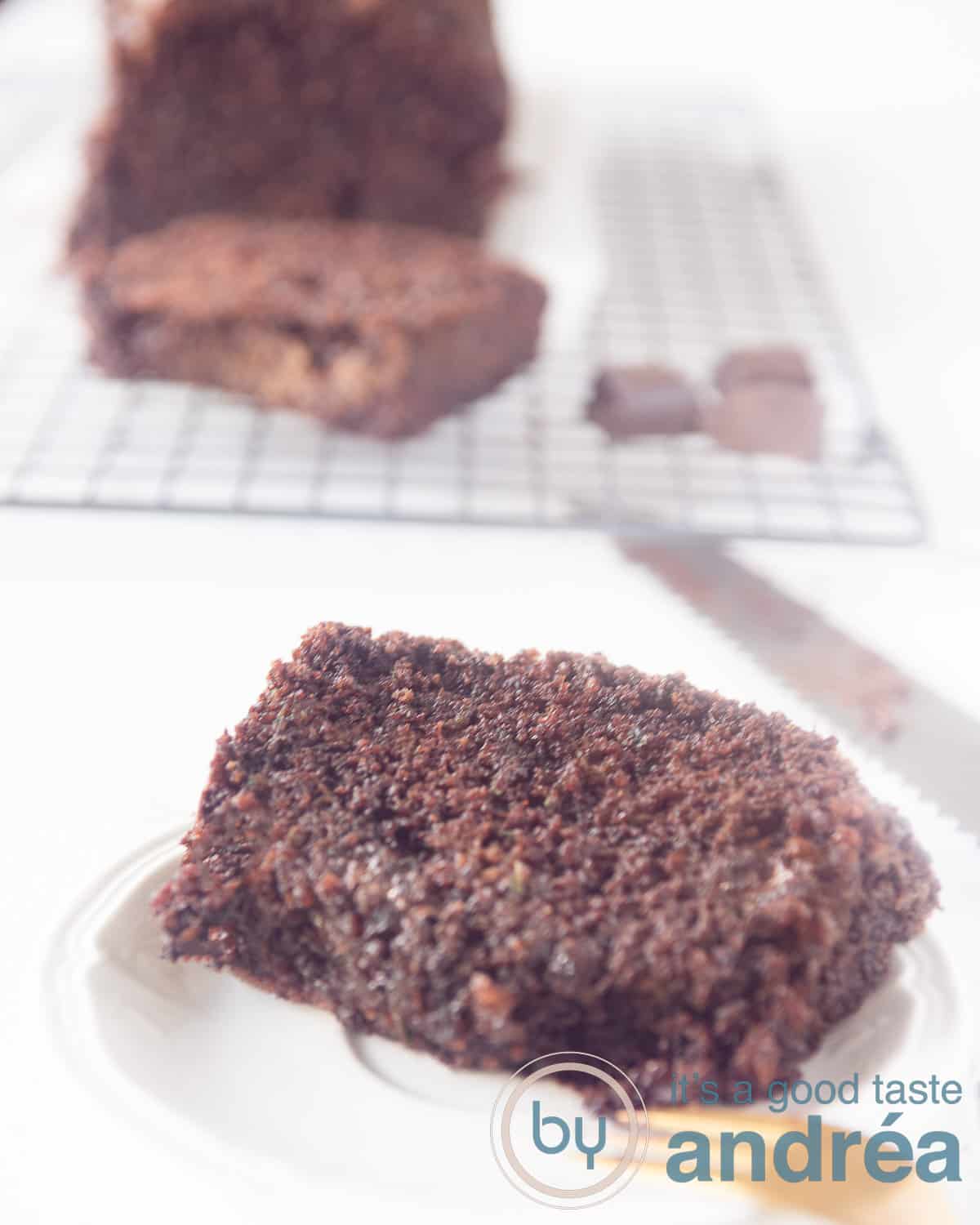 Een hoogtefoto met een wit bord met een plak courgettecake met chocolade ervoor. Achterin het resterende brood op een rek.