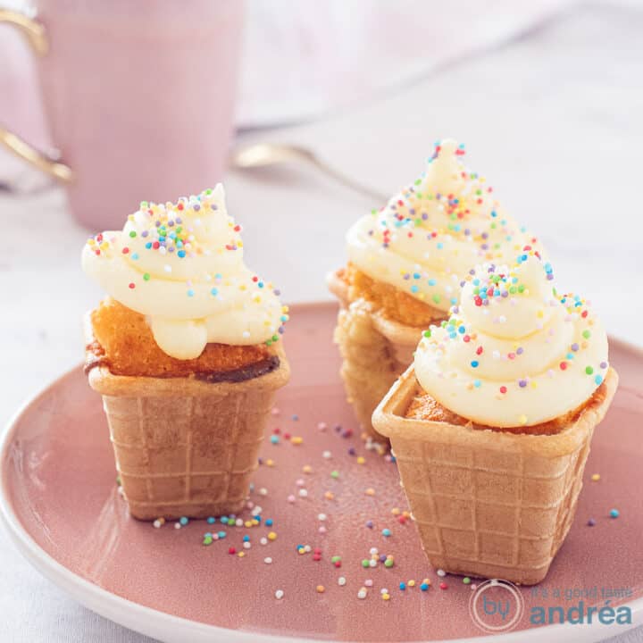 Een vierkante foto van 3 ijsbakjes van cupcake met frosting op een roze bord.