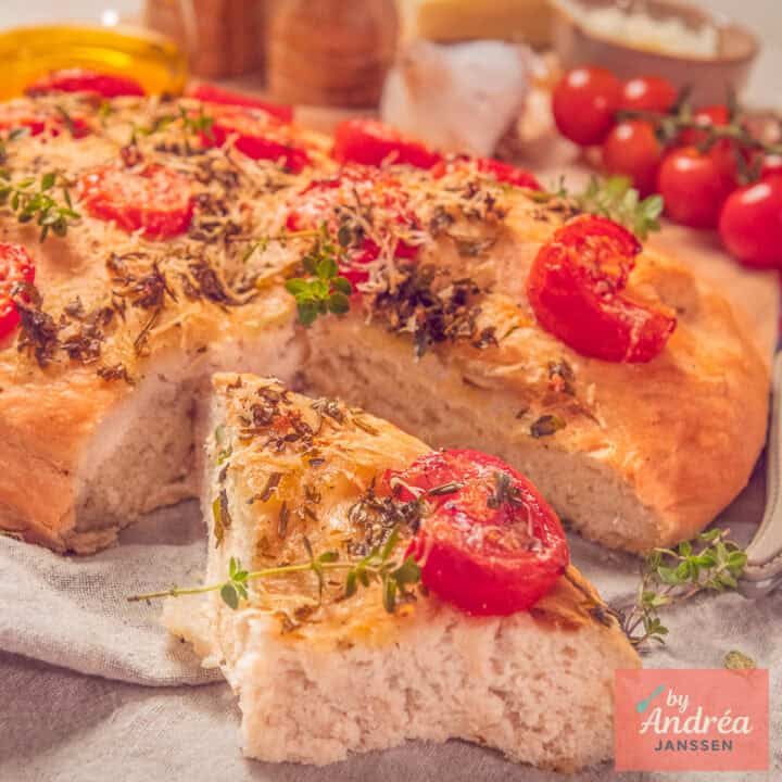 Een vierkante foto met een deel van een focaccia waar een punt uit is gesneden (waardoor je de luchtige binnenkant ziet). Met een topping van Italiaanse kruiden, tomaat, knoflook en Parmezaanse kaas.