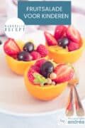 3 uitgeholde sinaasappels met druiven, bessen, frambozen en aardbeien. Bovenin een tekst: makkelijk recept, fruitsalade voor kinderen