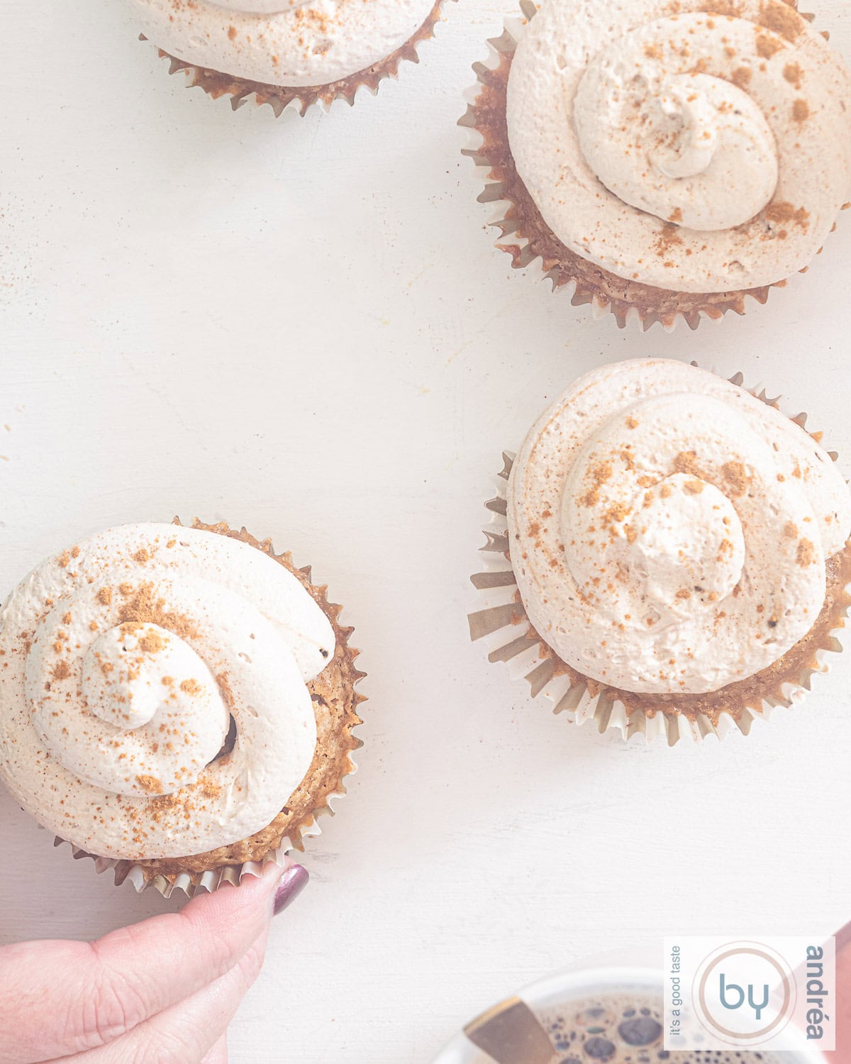 Een foto van bovenaf van vier cupcakes met een topping in tiramisu stijl. Een hand pakt een cupcake. Een witte ondergrond.