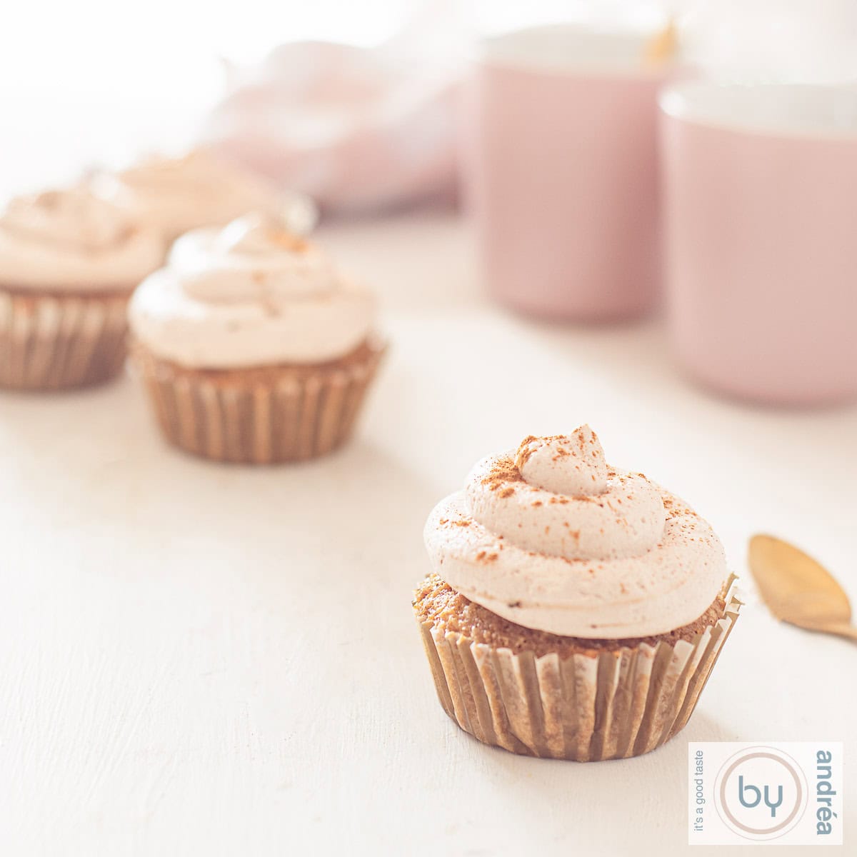 Een vierkante foto met een aantal cupcakes met tiramisu op een witte ondergrond. Aan de rechterzijde twee roze kopjes.