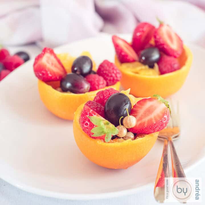 Een vierkante foto met drie uitgeholde sinaasappels gevuld met zomers fruit