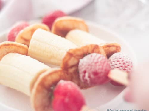 Een vierkante foto met drie stokjes gevuld met banaan, poffertjes en aardbeien. Een hand pakt er een van het bord.