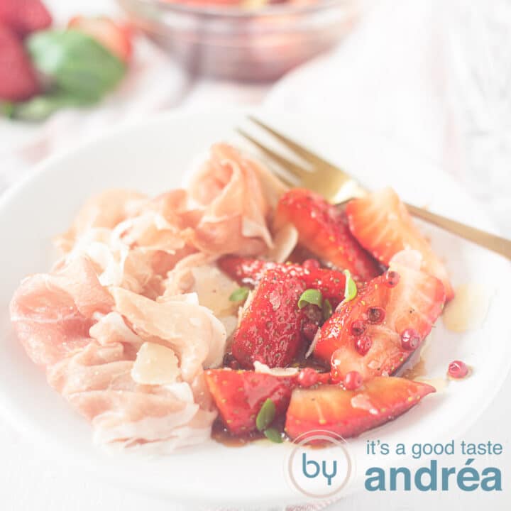 Een vierkante foto met een wit bord gevuld met balsamico aardbeien, ham, een gouden vork en in de achtergrond de ingrediënten