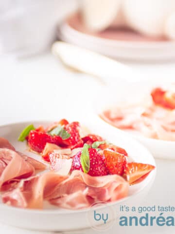 Op een wit bord aardbeien gemarineerd in creme balsamico met ham, op de achtergrond servies