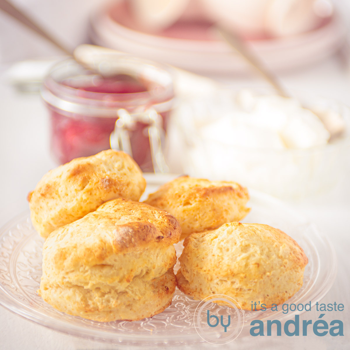Een vierkante foto met een glazen bord met scones en in de achtergrond een pot aardbeienconfituur.