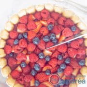Een vierkante foto van een charlotte russe van bovenaf. Een topping van frambozen, blauwe bessen en aardbeien. Een mes snijdt een partje eraf.