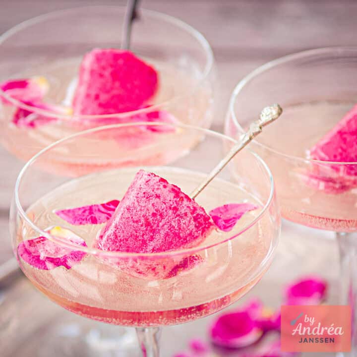 Een vierkante foto met drie champagneglazen met bubbels, frambozen ijscollies en rozenblaadjes