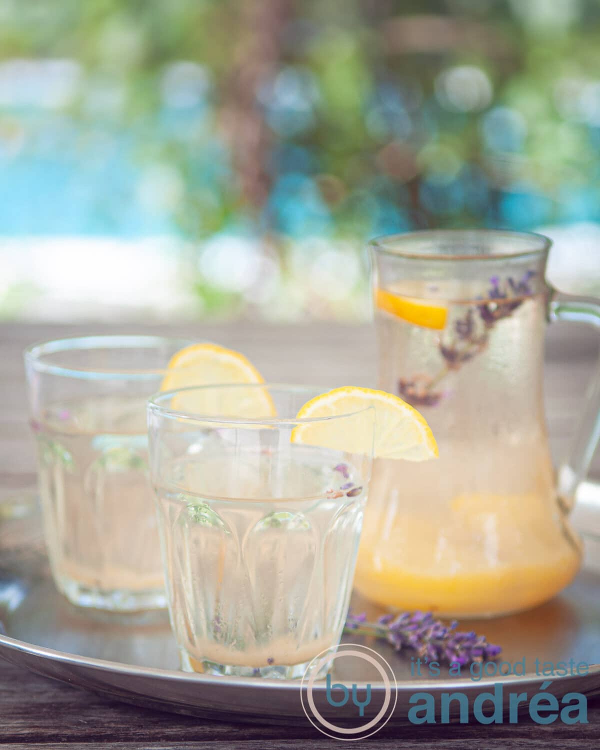 Twee glazen met lavendel limonade gegarneerd met citroen en een kan met limonade in een zomerse achtergrond