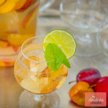Een vierkante foto met een glas met sangria met cava en zomerfruit. Een kan erachter.