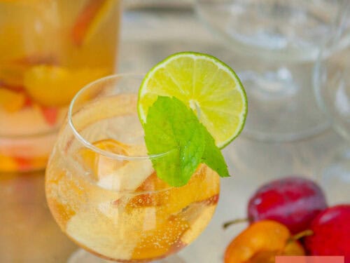 Een vierkante foto met een glas met sangria met cava en zomerfruit. Een kan erachter.