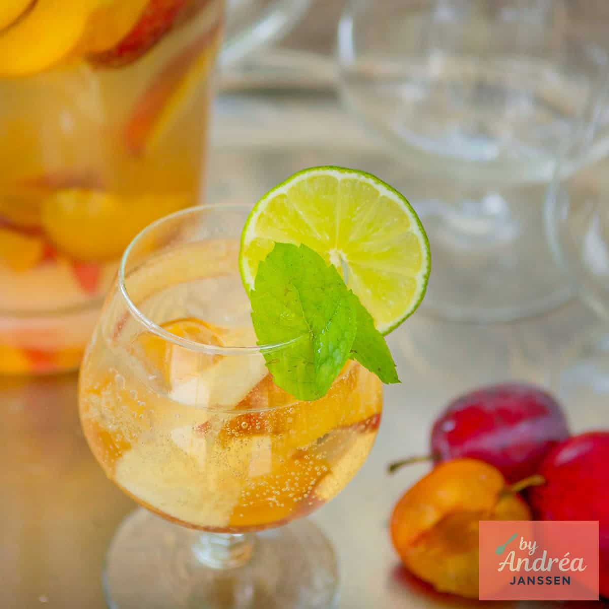 Een vierkante foto met een glas met sangria met cava en zomerfruit. Een kan erachter.