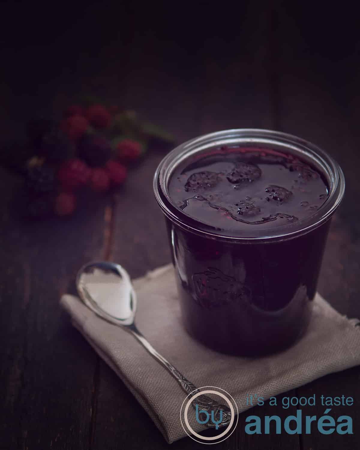 Een pot met Bramenjam met oregano op een bruine lap met een lepel