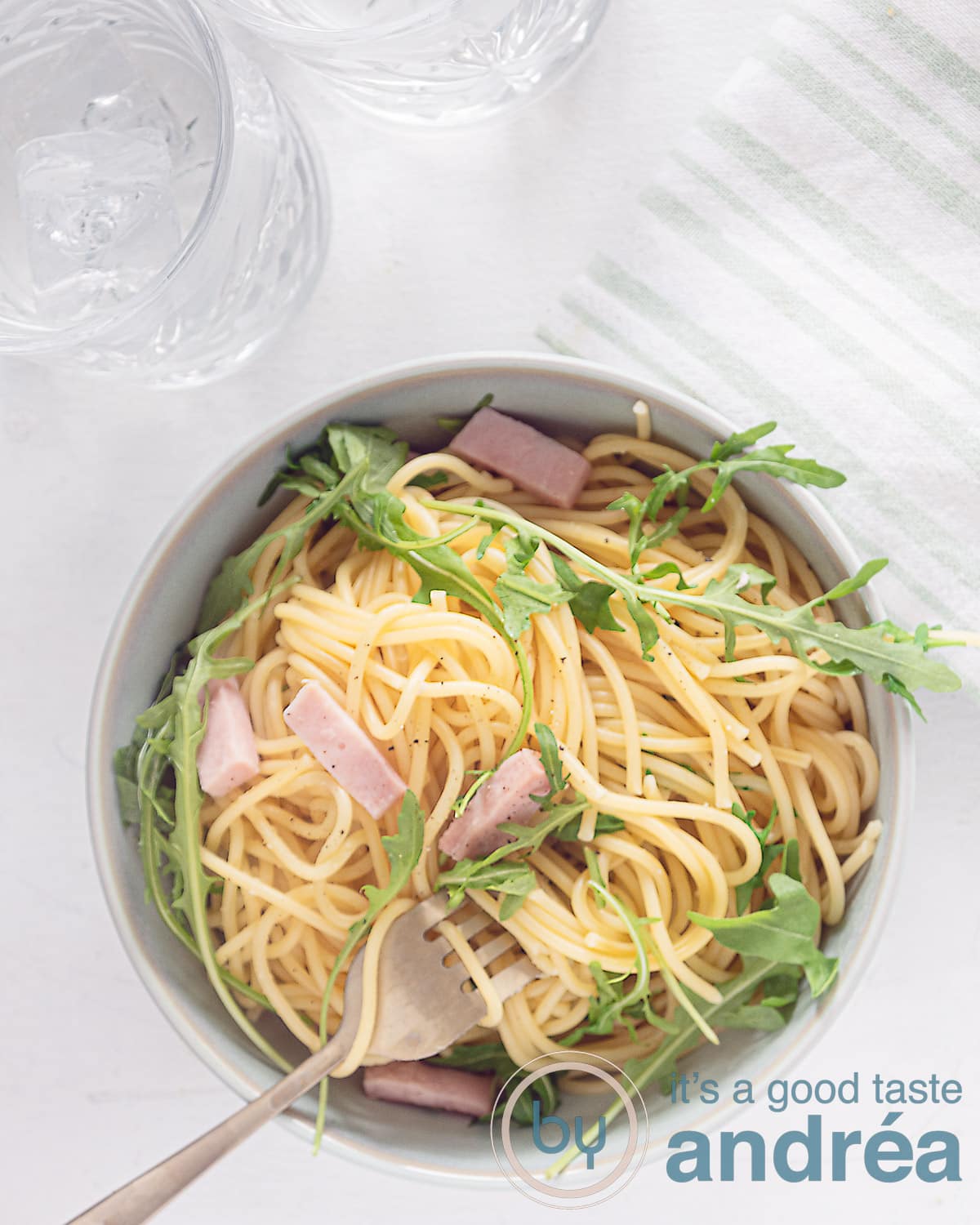 A photo from above of a white background with a bowl filled with spaghetti, ham and arugula. A golden fork with spaghetti curled up.