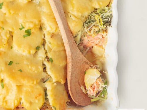 Een vierkante foto met een deel van een ovenschaal gevuld met zalm, prei en aardappelschijfjes