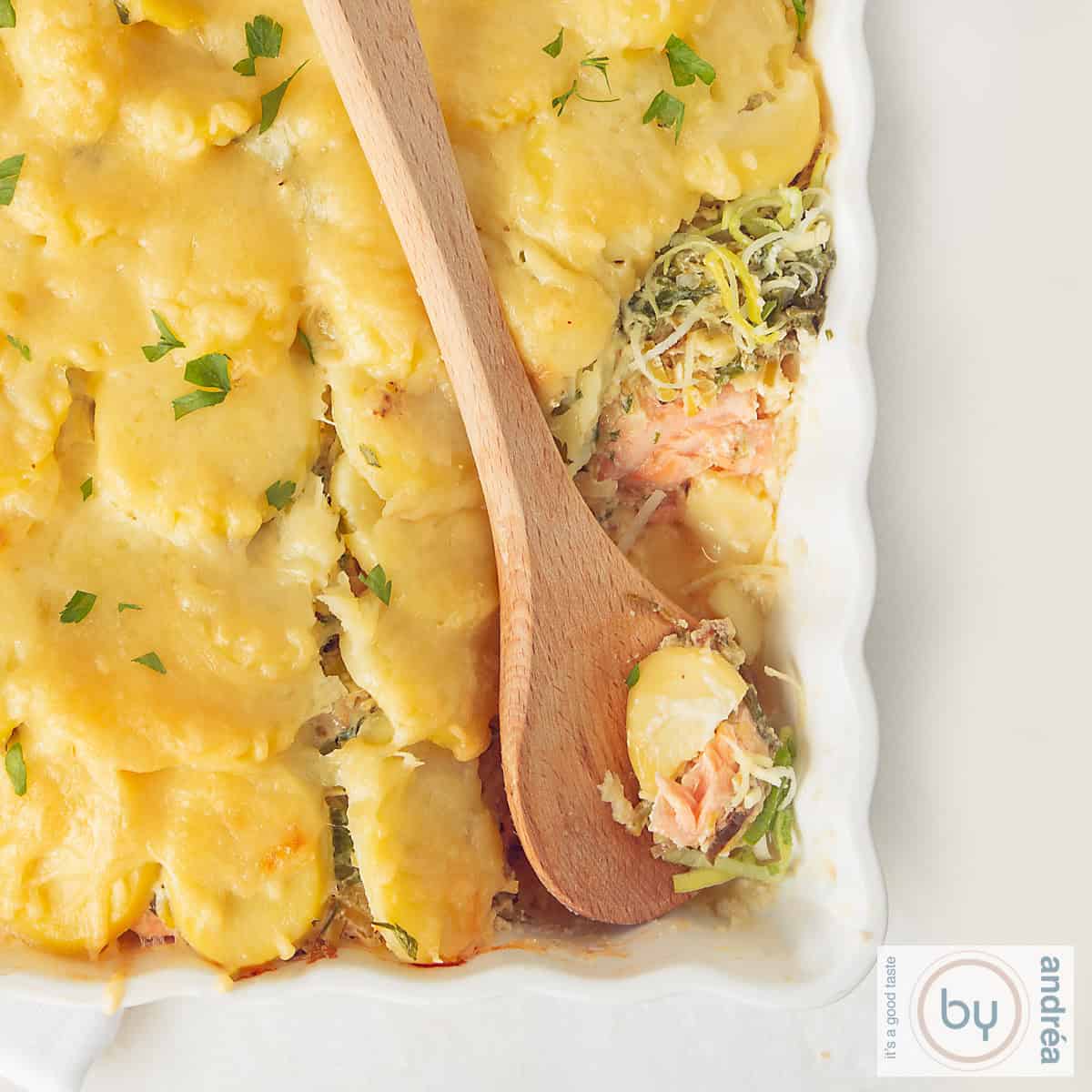 Een vierkante foto met een deel van een ovenschaal gevuld met zalm, prei en aardappelschijfjes