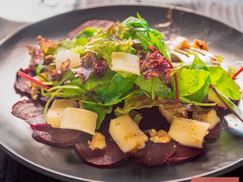 Een vierkante foto met een bord met carpaccio van bieten, peer, kaas, sla en walnoten