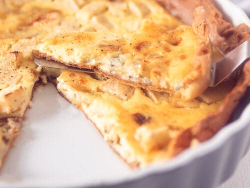 Een vierkante foto met een gorgonzola quiche met appel. Een punt eruit gesneden, die ligt er bovenop.