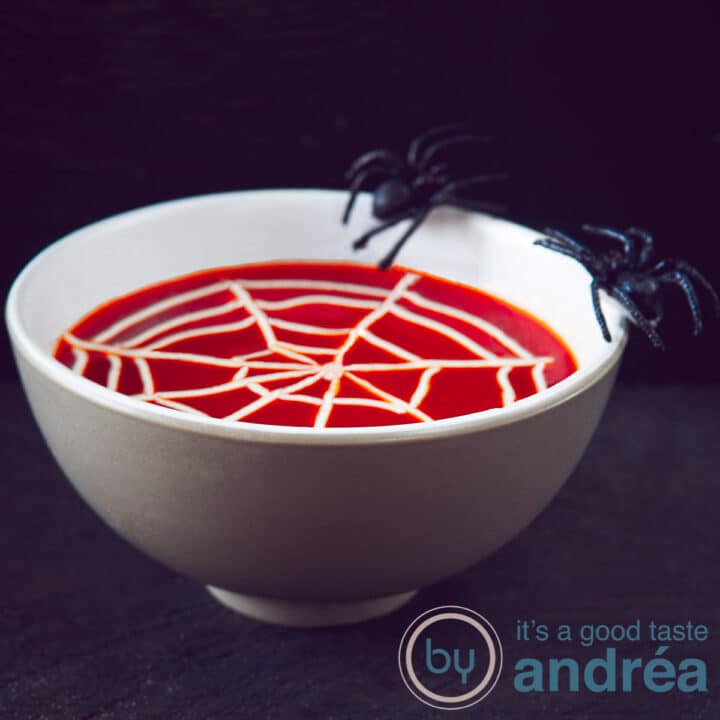 Een vierkante foto met een zwarte achtergrond een grijze kom gevuld met romige tomatensoep met een spinnenweb. Op de rand balanceren twee spinnen.