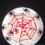 Een donkere achtergrond met een wit bord. Daarop een witte taart met een rood spinnenweb en twee M&M spinnen. Om de taart zwarte spinnen