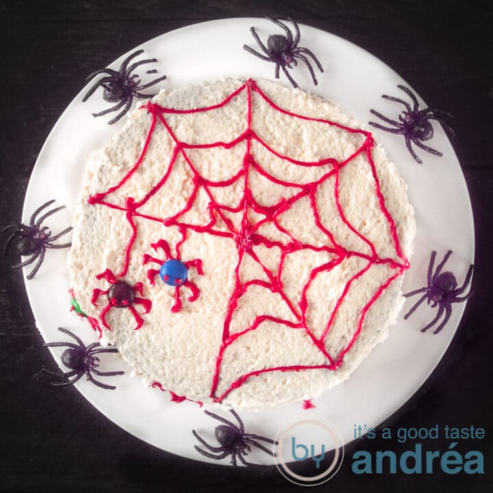 Een vierkante foto met een wit bord met een taart met rood spinnenweb en twee spinnen. Er omheen spinnen.
