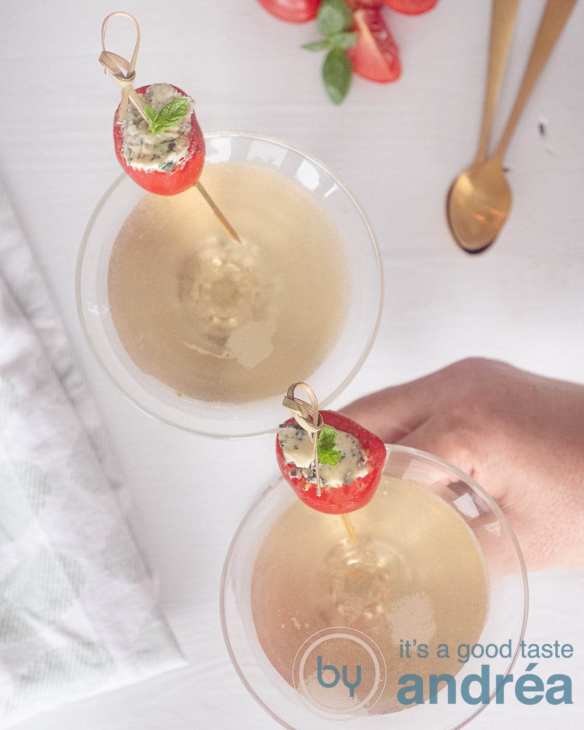 Een foto van bovenaf van twee glazen gevuld met martini met Gin en Vermout en een cherry tomaatje als garnering. Een hand pakt een glas. Een witte ondergrond