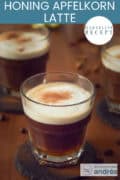 Een bruine tafel met daarop drie kopjes koffie met melkschuim. Een tekst bovenin: makkelijk recept, honing apfelkorn latte