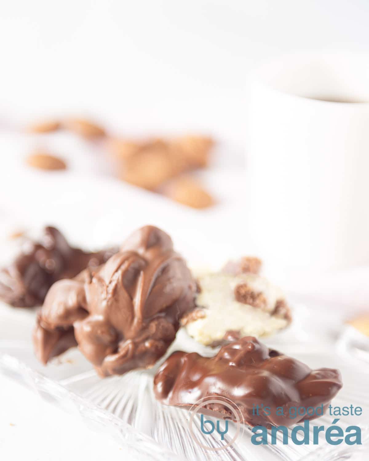 Een witte achtergrond. Voorin een glazen bord met drie verschillende soorten pepernoten chocolaatjes. Een witte mok en wat pepernoten achterin.