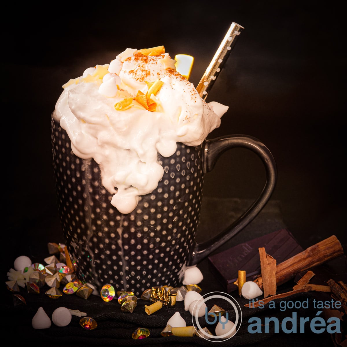 Een vierkante foto van een zwarte mok met slagroom, gouden chocolade krullen en op de zwarte ondergrond kaneel, marshmallows.