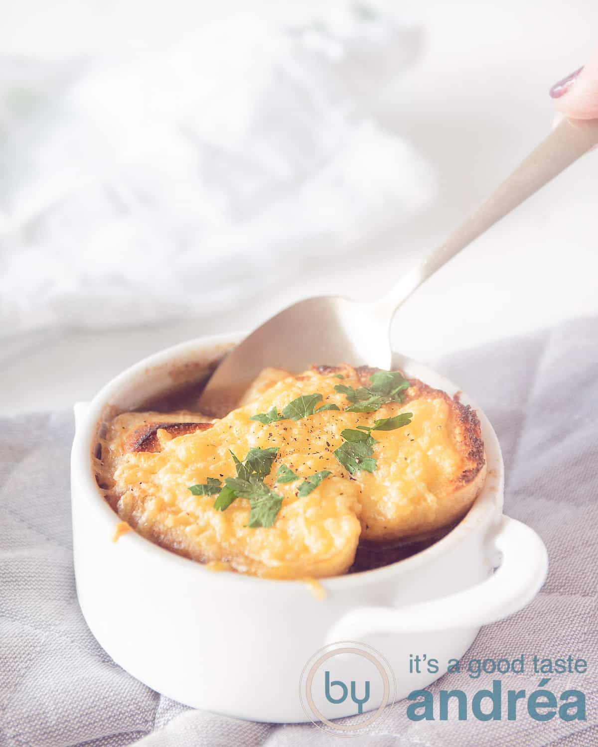 Een witte kom met Franse Uiensoep met twee stukjes stokbrood gegratineerd. Een gouden lepel neemt een hap van de soep