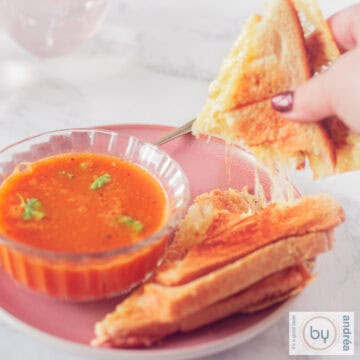 Een vierkante foto met een kom met tomatensoep met peterselie en een tosti waar een stuk is afgebroken waardoor slierten gesmolten kaas zichtbaar zijn