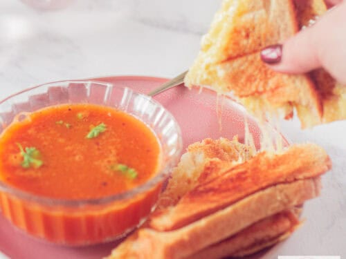 Een vierkante foto met een kom met tomatensoep met peterselie en een tosti waar een stuk is afgebroken waardoor slierten gesmolten kaas zichtbaar zijn