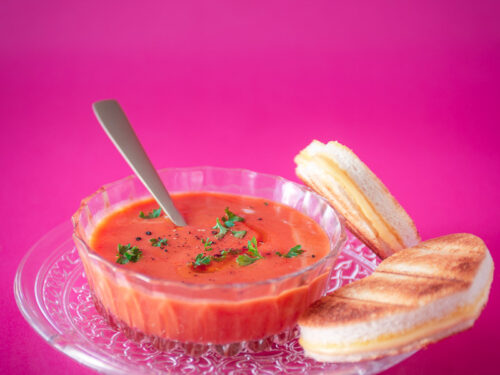 een glazen kom met tomatensoep, hartjes tosti op een roze achtergrond