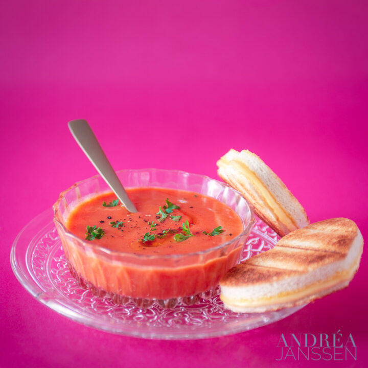 een glazen kom met tomatensoep, hartjes tosti op een roze achtergrond