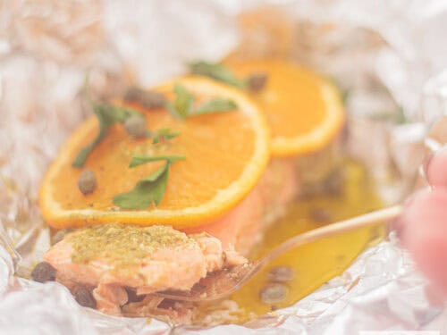Een vierkante foto met aluminiumfolie, waarin gesmolten boter, zalm, sinaasappel, pesto en kappertjes zijn gegaard.