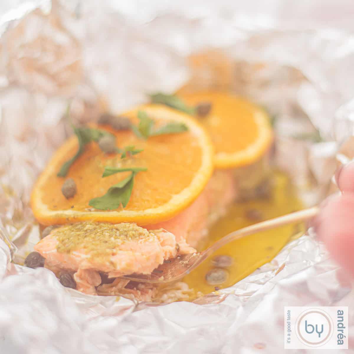 Een vierkante foto met aluminiumfolie, waarin gesmolten boter, zalm, sinaasappel, pesto en kappertjes zijn gegaard.