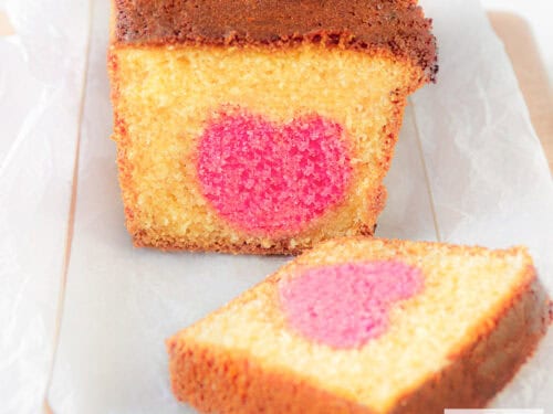 Een vierkante foto met een glazen schaal met daarop een cake waar een plakje is afgesneden. Een roze hart kun je zien